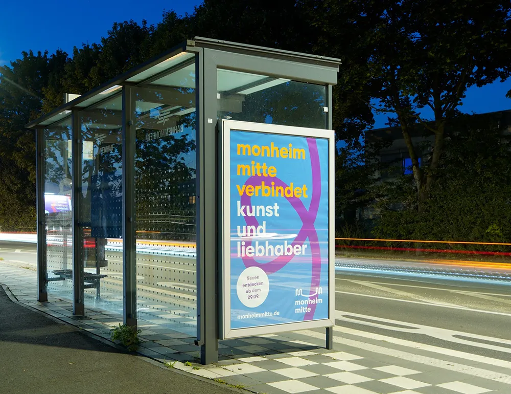 Plakat der Kampagne Monheim Mitte auf einem Buswartehäuschen in einer abendlichen Außenanlage. Titel auf Plakat: monheim mitte verbindet kunst und liebhaber