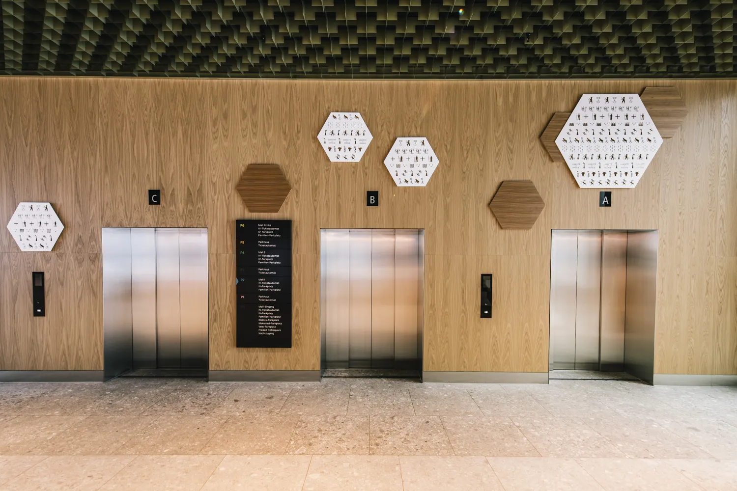 Frontale Ansicht von drei Aufzügen und einem Wegeleitsystem in einem Vorraum, dessen Wände aus Holzvertäfelungen und grafischen sechseckigen Elementen bestehen
