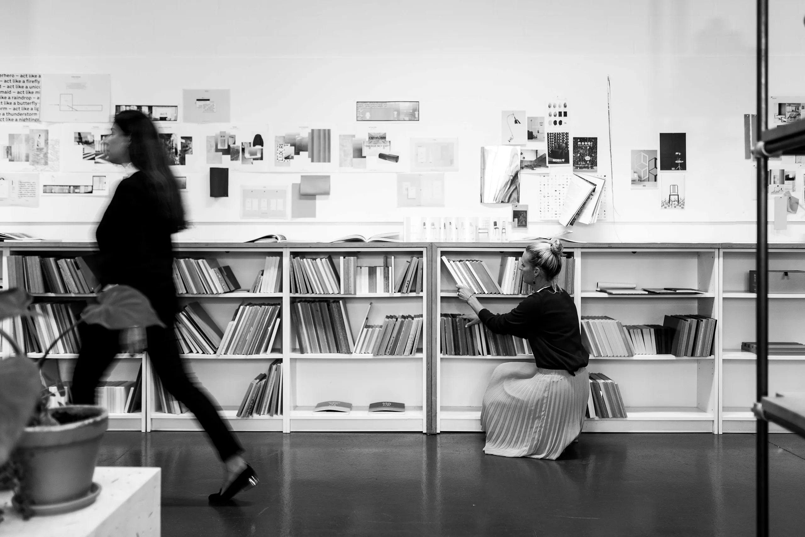 Zwei Mitarbeiterinnen im Büro vor Bücherregalen und Moodboard. Eine im Vorbeigehen, die andere kniet und holt ein Buch raus.