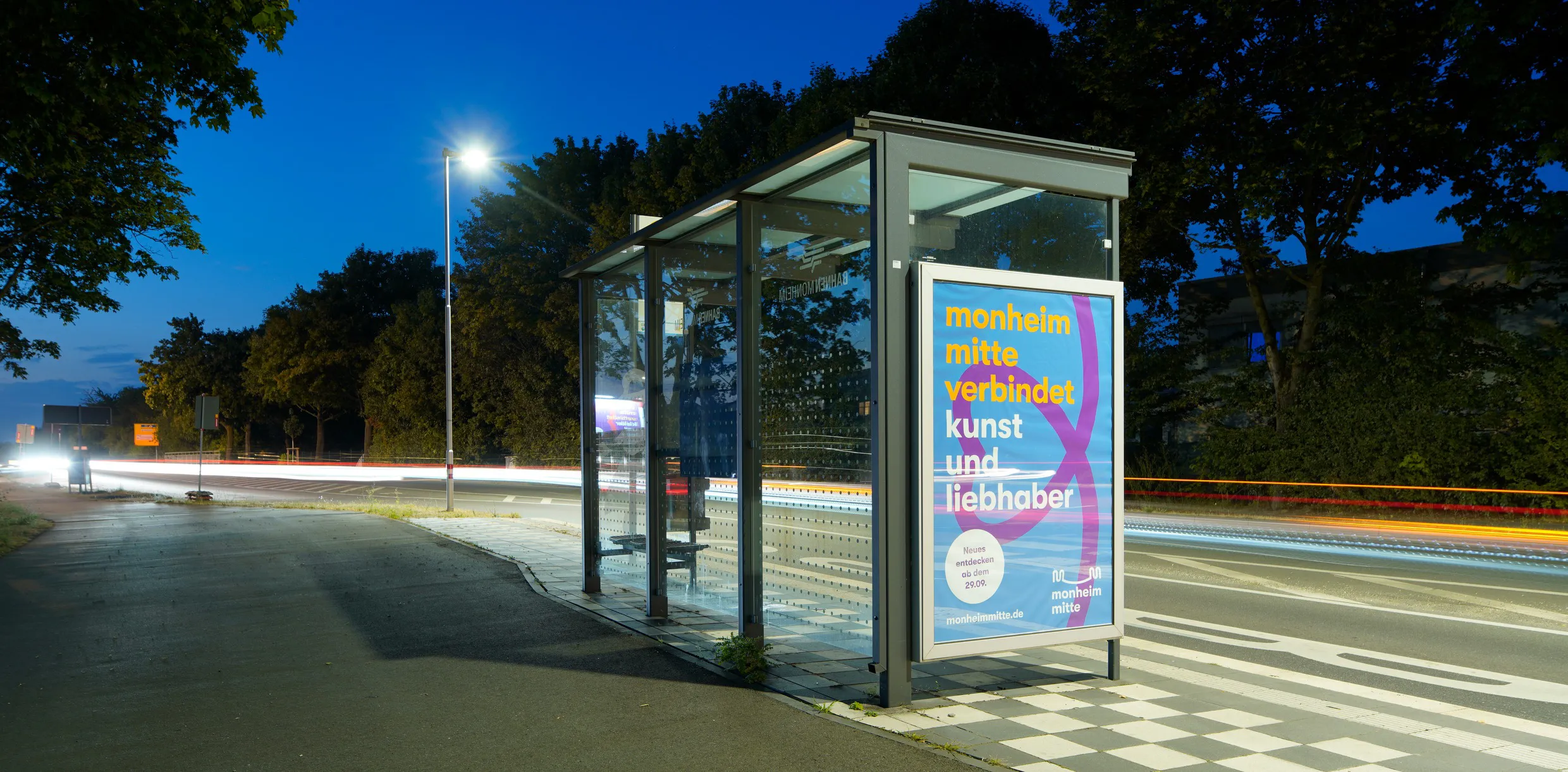 Plakat der Kampagne Monheim Mitte auf einem Buswartehäuschen in einer abendlichen Außenanlage. Titel auf Plakat: monheim mitte verbindet kunst und liebhaber