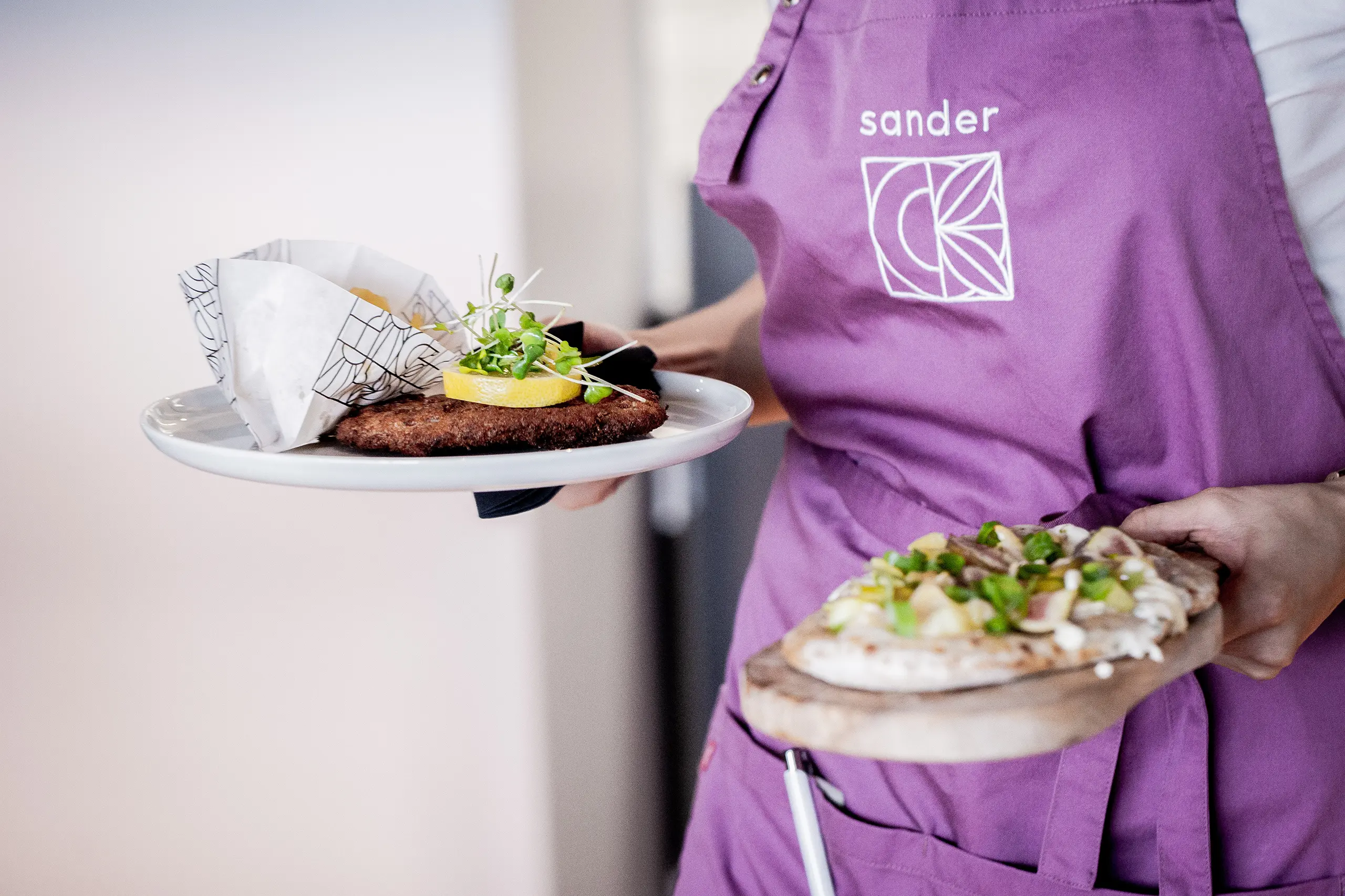 Bild eines Kellners, der eine violette Schürze mit dem neuen Sander-Logo trägt, während er zwei Teller mit Speisen hält.